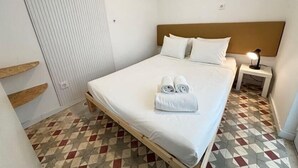 Una cama individual con estructura de madera, ropa de cama blanca y cabecera. Hay una mesita de noche pequeña con una lámpara, y un suelo con estampado geométrico.