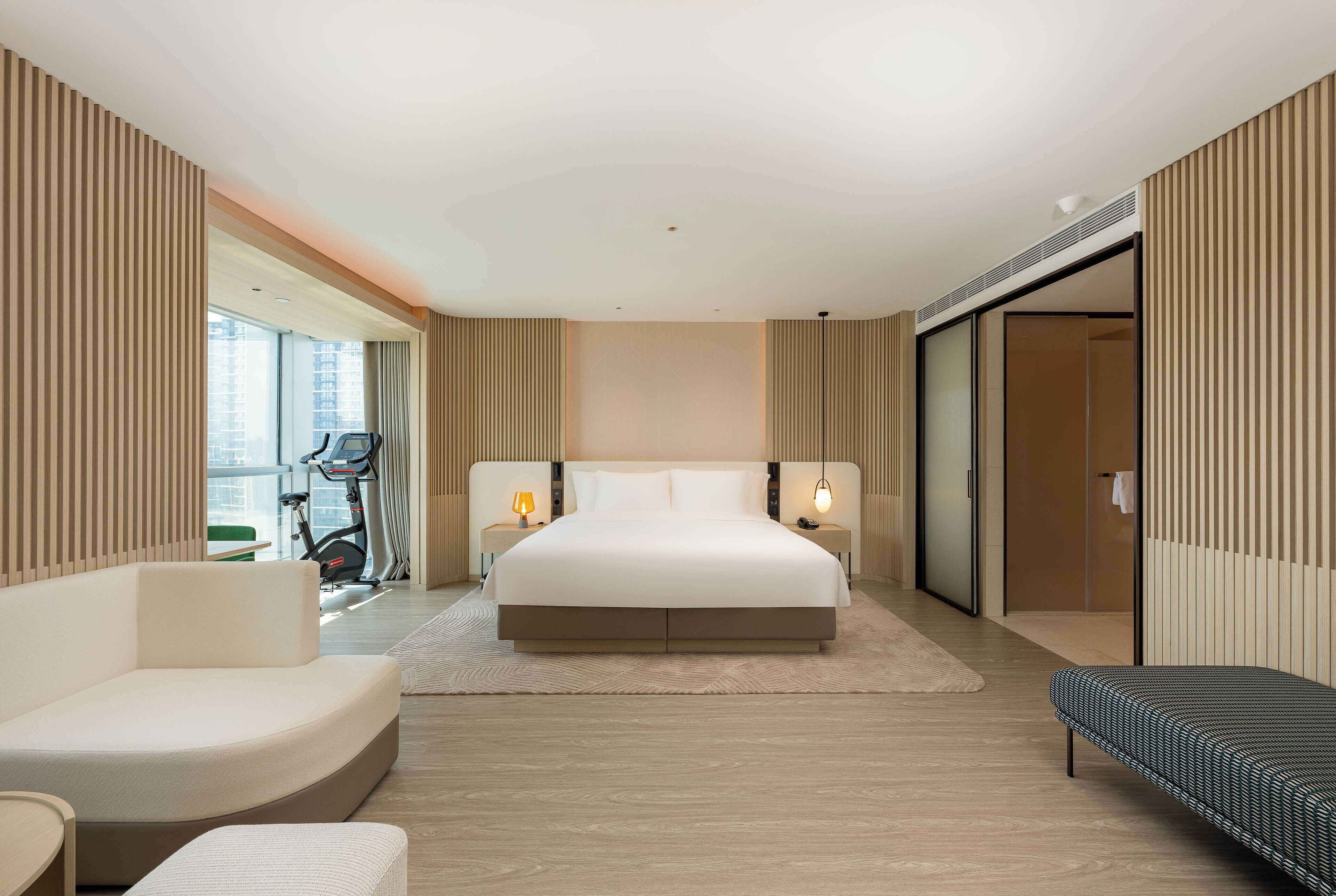 A modern hotel room with a large bed, a sofa, a treadmill, and a sliding door closet.