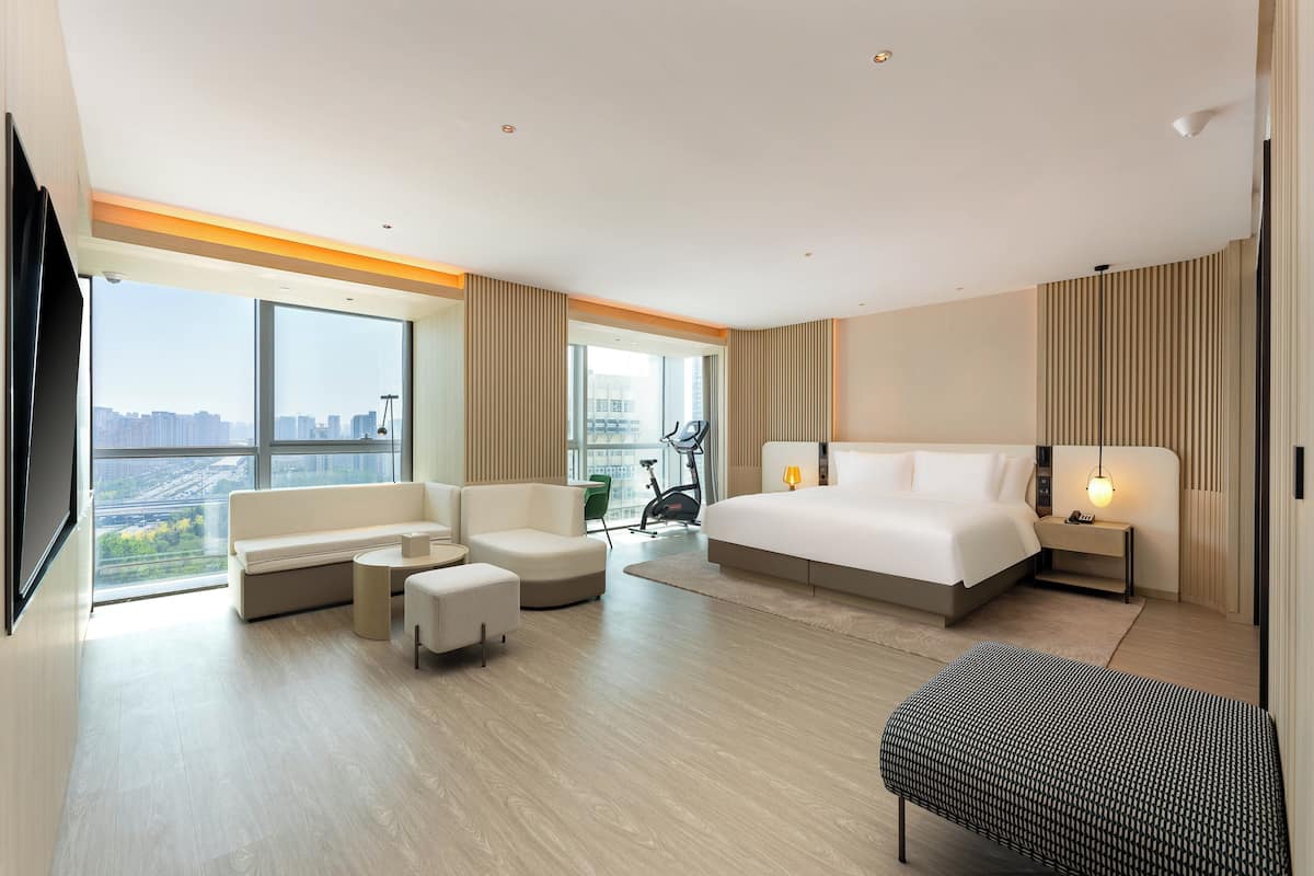 Habitación de hotel moderna con una cama grande, un sofá, una mesita y una silla. Se ve una ventana con vista a la ciudad y una bicicleta en la esquina.