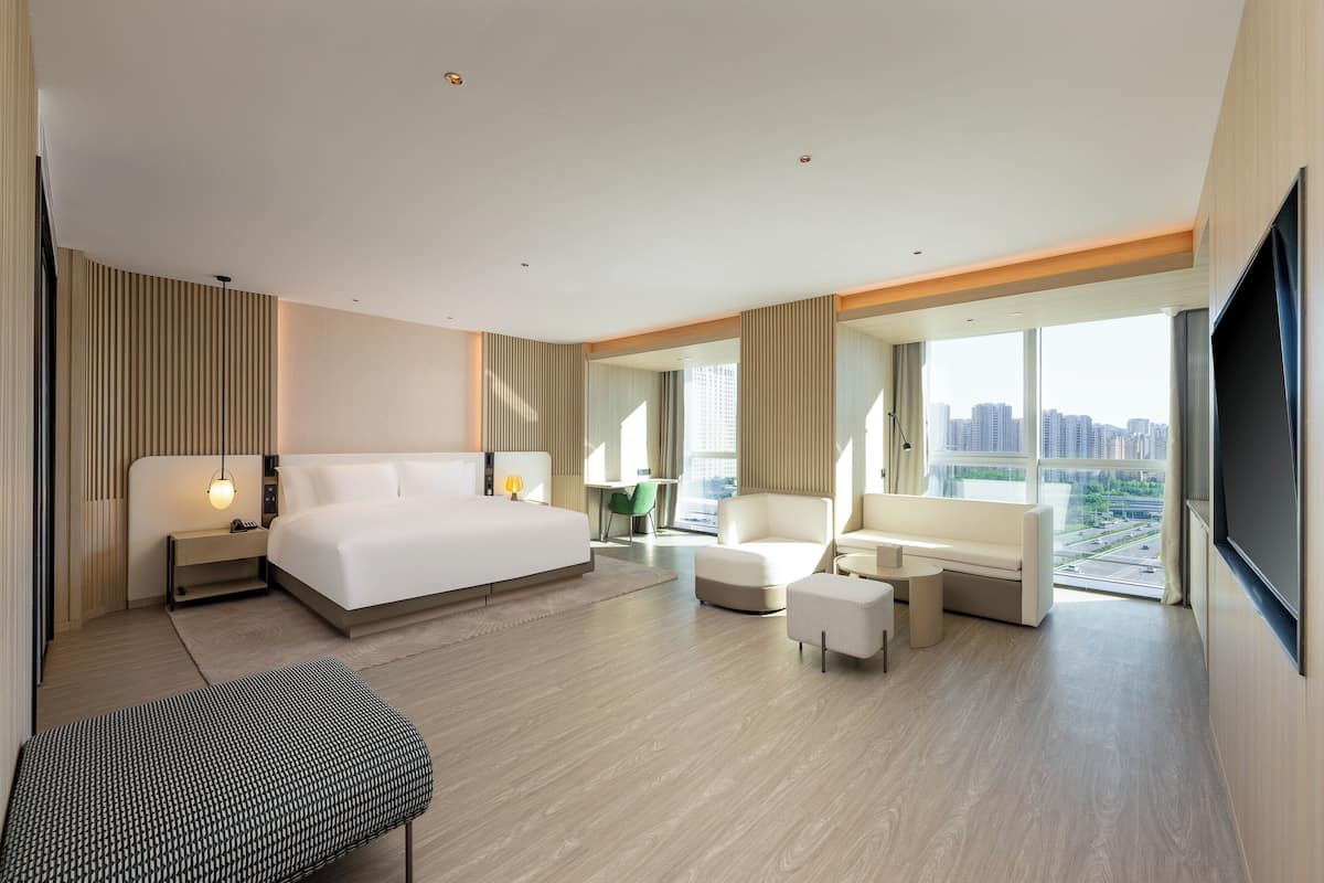 Una habitación de hotel moderna con una cama grande, una zona de estar con sofá y sillones, una mesita y un ventanal con vistas a la ciudad.
