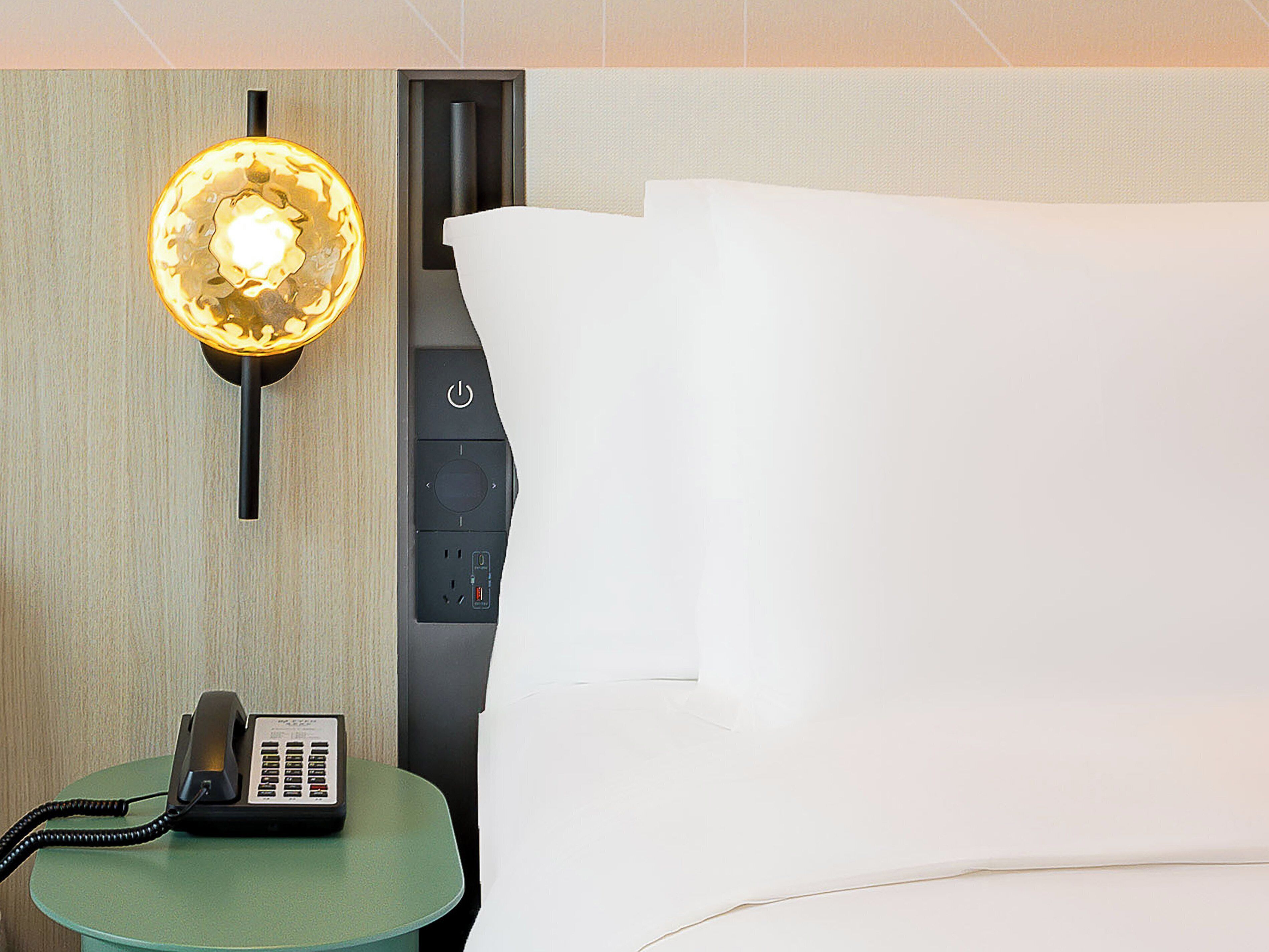 A hotel room with a bed, a bedside table with a telephone, and a wall-mounted light fixture.