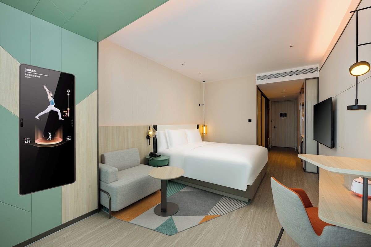 Una habitación de hotel moderna con una cama, un sofá, una mesita, un televisor y una pantalla digital mostrando una postura de yoga.
