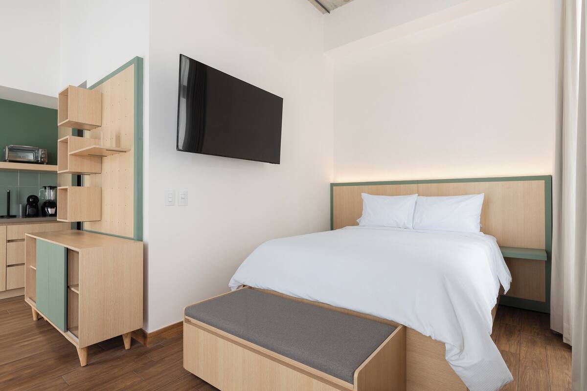 A modern hotel room with a bed, a TV mounted on the wall, a wooden desk with a shelf, and a bench.