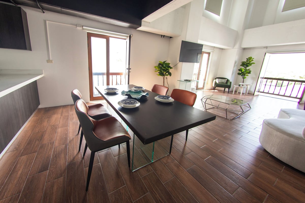 A modern dining area with a black table, brown chairs, and a glass coffee table. There are two potted plants and a flat-screen TV mounted on the wall.