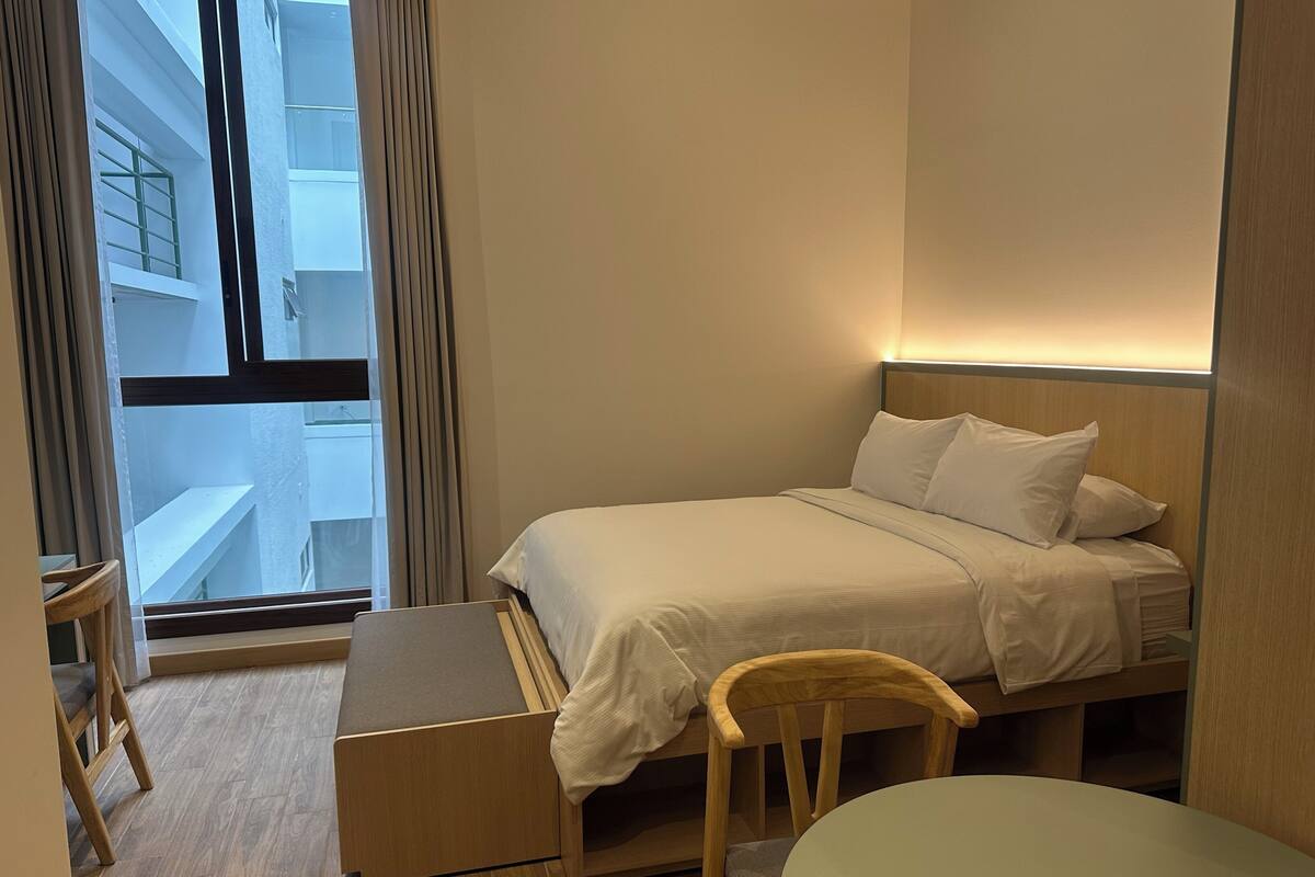 A hotel room with a bed, bedside table, chair, and a view of a building through a window.