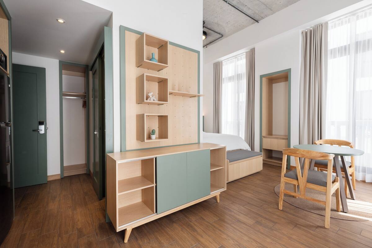 A modern hotel room with a wooden floor, a bed with a green headboard, a built-in wooden shelf unit, and a dining area with a table and chairs.