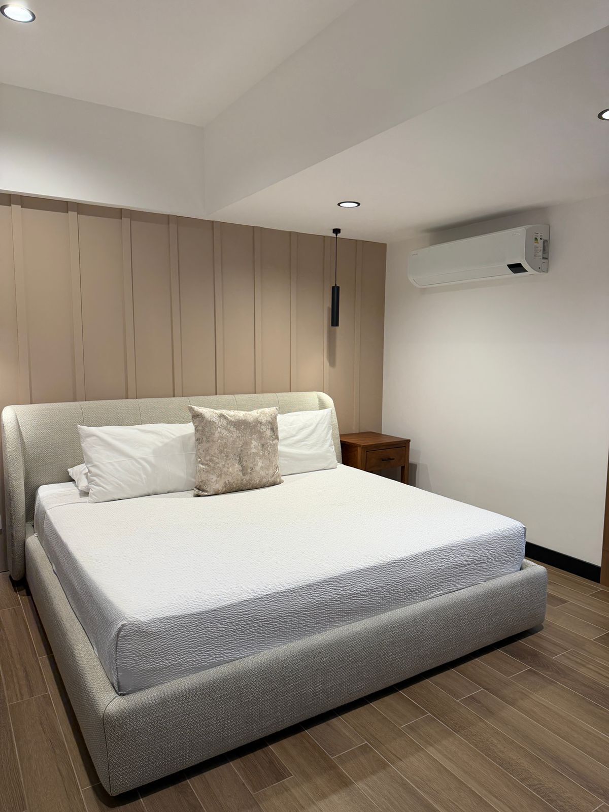 A modern bedroom with a large bed, a bedside table, a wall-mounted air conditioner, and a ceiling light.