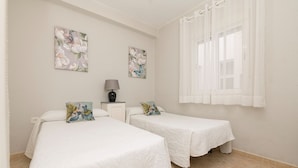 A room with two beds, each with a white bedspread, a small bedside table with a lamp, and two floral paintings on the wall. - PLAYAMAR - Nice flat near the sea in Cadiz and Free WiFi (Cádiz)