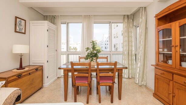 A dining area with a wooden table and chairs, a plant centerpiece, a wooden cabinet, and a view of buildings through large windows. - PLAYAMAR - Nice flat near the sea in Cadiz and Free WiFi (Cádiz)