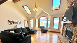 Un salon spacieux avec un canapé d’angle, un fauteuil inclinable et une cheminée en pierre. De grandes fenêtres offrent une vue sur l’extérieur.