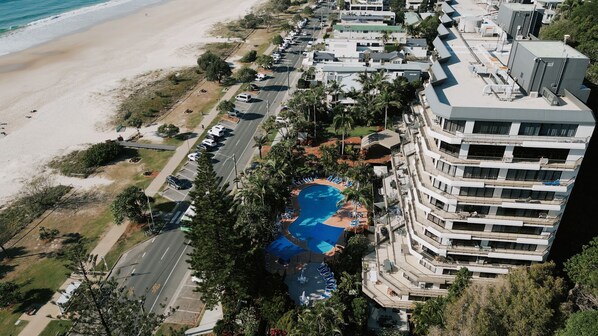 Vista aérea de un hotel frente a la playa con piscina, varios balcones y un camino bordeando la playa.