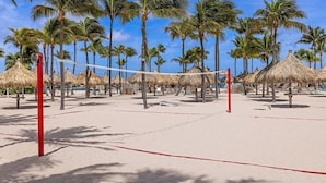 A beach volleyball court with palm trees and thatched umbrellas.