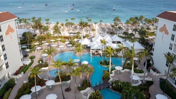 A resort with a central swimming pool, palm trees, and a view of the ocean. - Palm Beach Aruba | Couples Caribbean Getaway | Pool + Beach (Palm Beach)