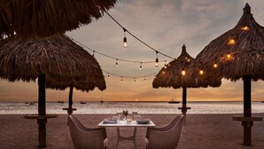 A beach dining setup with thatched umbrellas, string lights, and a table with chairs. - Palm Beach Aruba | Couples Caribbean Getaway | Pool + Beach (Palm Beach)