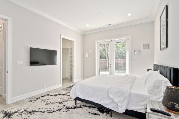A modern bedroom with a large bed, a flat-screen TV mounted on the wall, and a sliding glass door leading to a balcony.