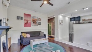 Una sala de estar moderna con sofá, mesa de centro y ventilador de techo. Se observa una zona de cocina con armarios y refrigerador.