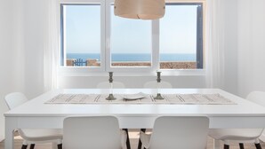A dining area with a white table and chairs, a wooden pendant light, and a view of the sea through large windows.