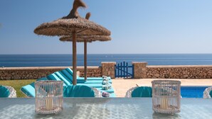 A seaside terrace with a thatched umbrella, lounge chairs, a stone wall, and a view of the ocean.