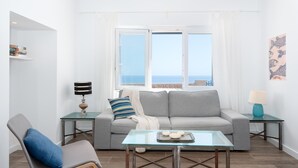 A modern living room with a grey sofa, a glass coffee table, and a blue chair. There are two side tables with lamps, and a view of the sea through large windows.