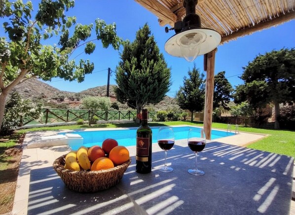 A poolside setting with a table, a bottle of wine, and a basket of fruit.