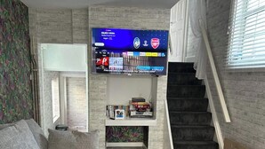 A living room with a flat-screen TV mounted on a brick wall, a staircase, and a door leading to another room. - The Hideaway ll /private garden/Quiet/Central (Southport)