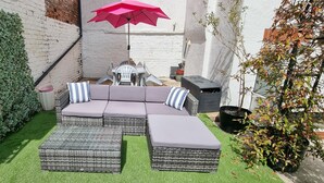 A cozy outdoor seating area with a wicker sofa and ottomans, a red umbrella, and a small table with chairs. - The Hideaway ll /private garden/Quiet/Central (Southport)