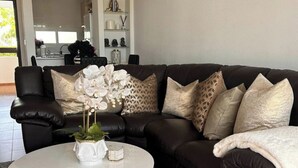 Un sofá de cuero marrón oscuro con almohadones decorativos y un jarrón blanco con orquídeas sobre una mesita redonda.