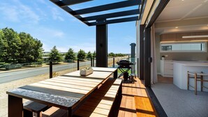 Un patio exterior moderno con mesa de madera y una parrilla, con vistas a una carretera y follaje.