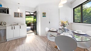 Una cocina moderna con armarios blancos, un microondas y un lavavajillas. Hay un comedor con una mesa de vidrio y sillas blancas. Un jarrón con flores está sobre la mesa.
