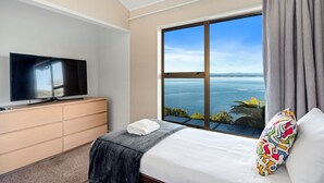 A hotel room with a large window offering a view of the sea, a bed with a white pillow, a flat-screen TV on a wooden dresser, and a grey curtain.