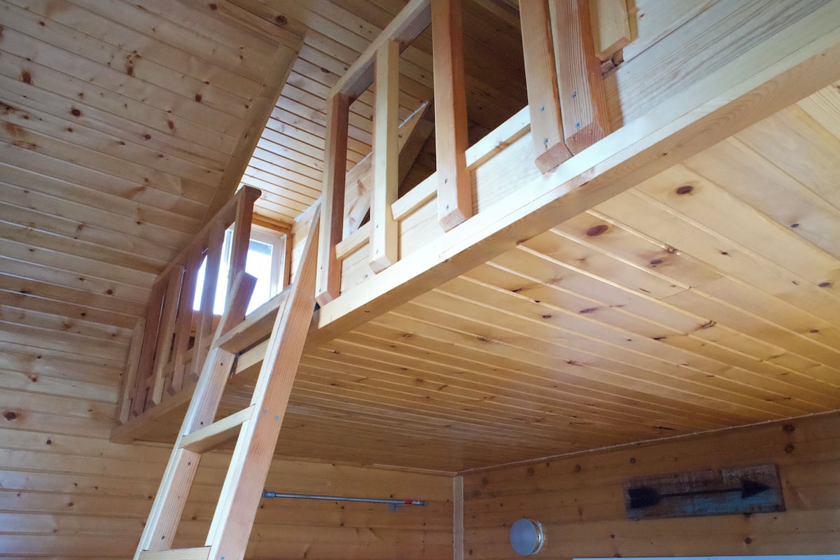 A wooden interior with a loft-style sleeping area, a ladder, and a visible window.