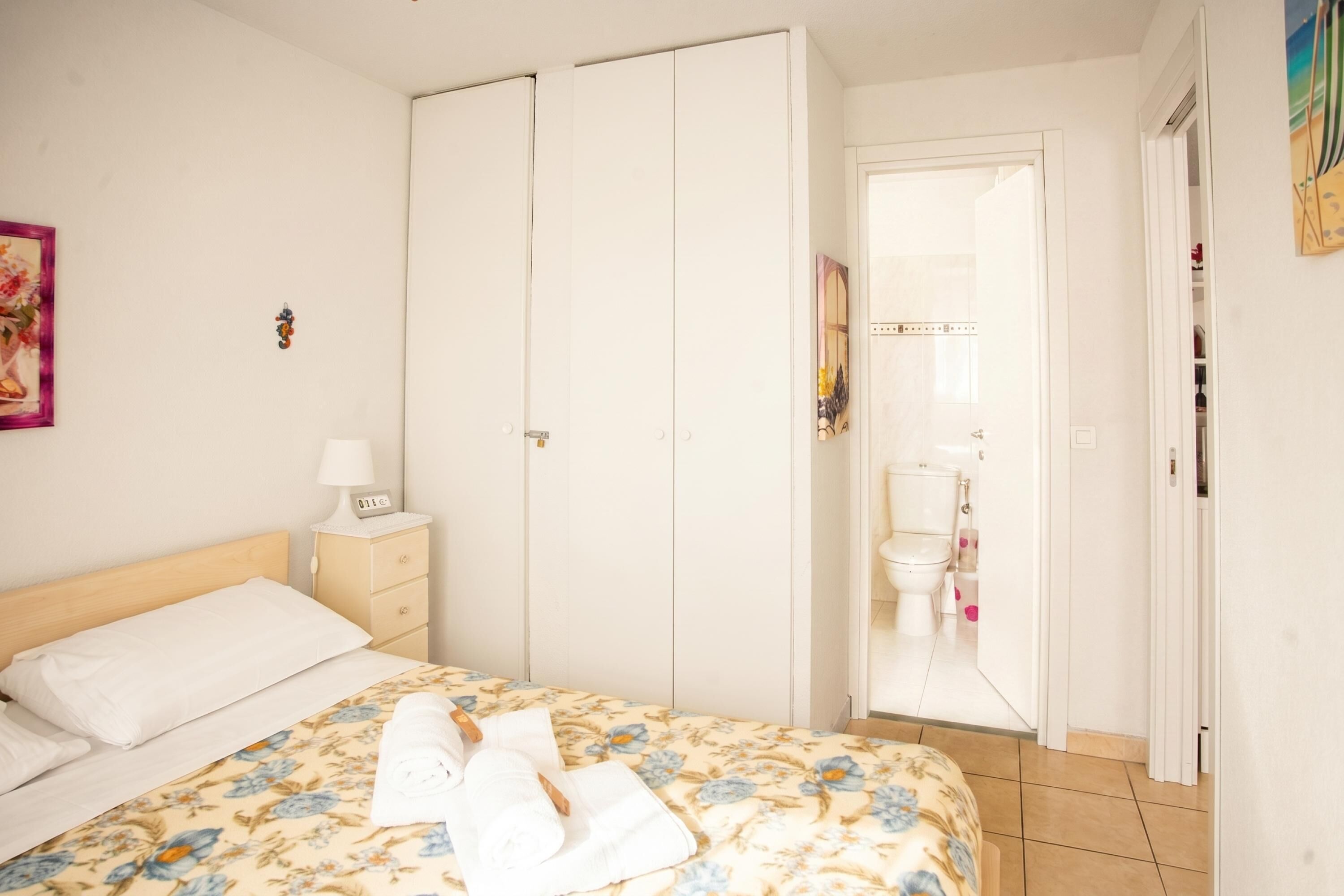 A bedroom with a bed, a nightstand, a wardrobe, and a bathroom visible through an open door.
