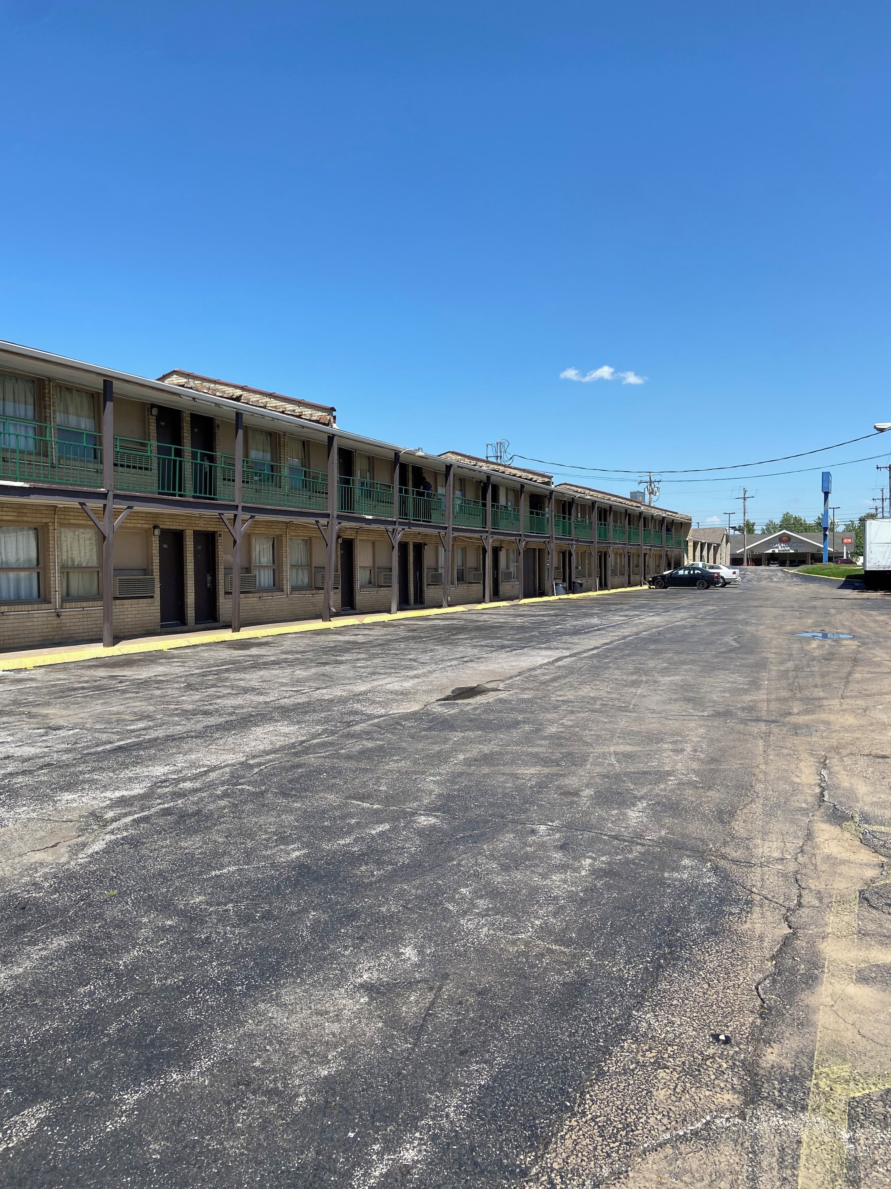 A row of two-story motel buildings with balconies, a parking lot, and a clear blue sky.