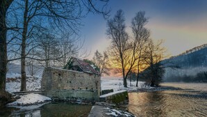 Un bâtiment rustique en pierre, avec un petit étang devant, entouré d’arbres sans feuilles et se fondant dans un paysage brumeux.