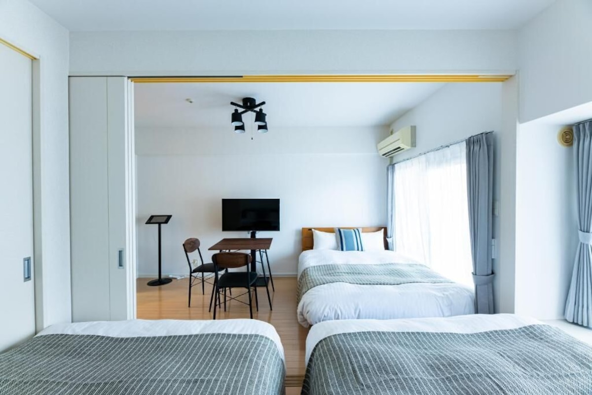 Ein Hotelzimmer mit zwei Betten, einem Schreibtisch mit Stuhl, einem Fernseher und einem Deckenventilator.