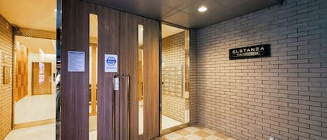 Un pasillo con una puerta de madera, un letrero que dice 'ELSTANZA TENJINHIGASHI' y un piso de baldosas.