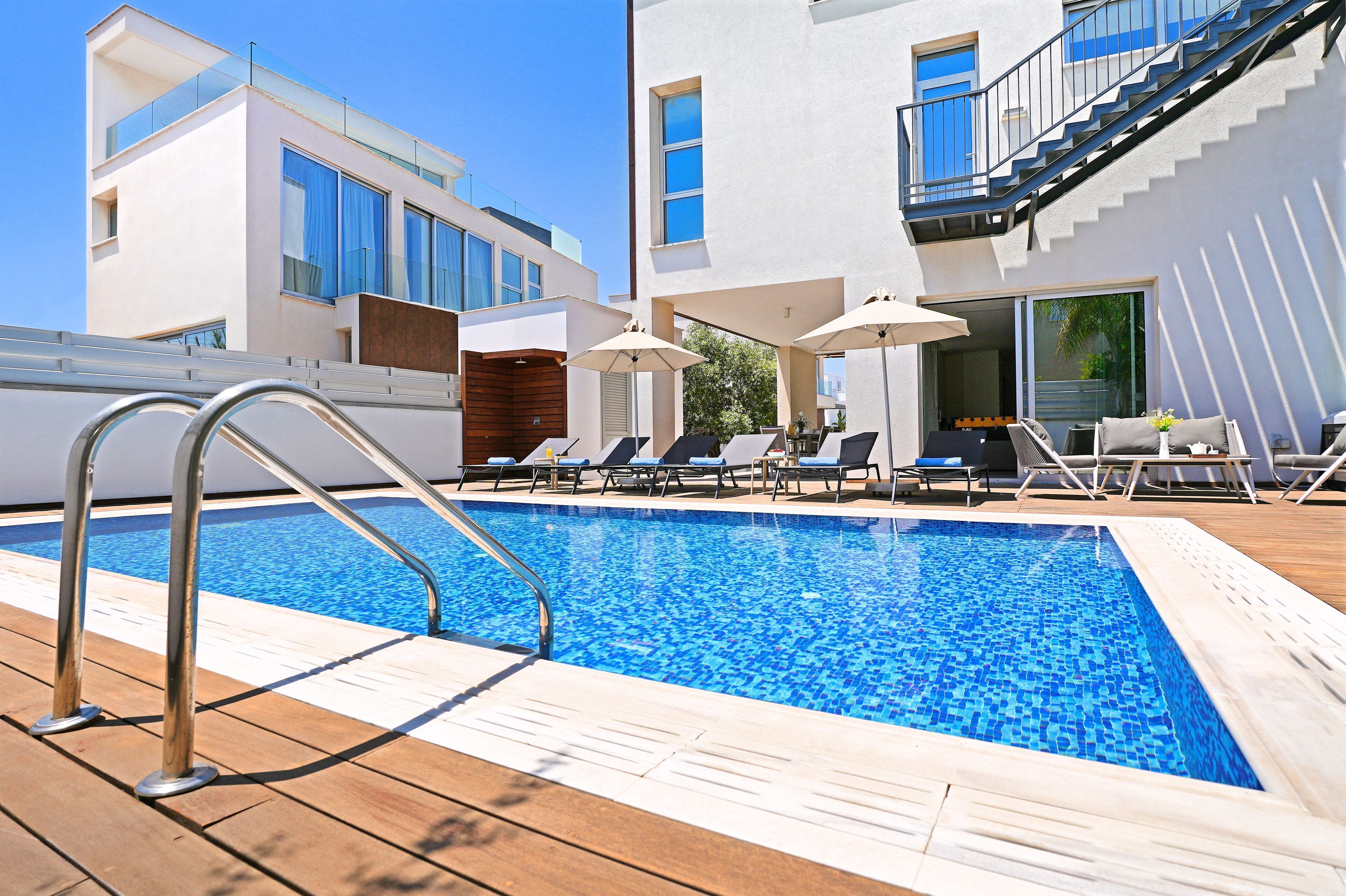 A swimming pool with sun loungers and umbrellas, surrounded by modern buildings with large windows and a staircase.