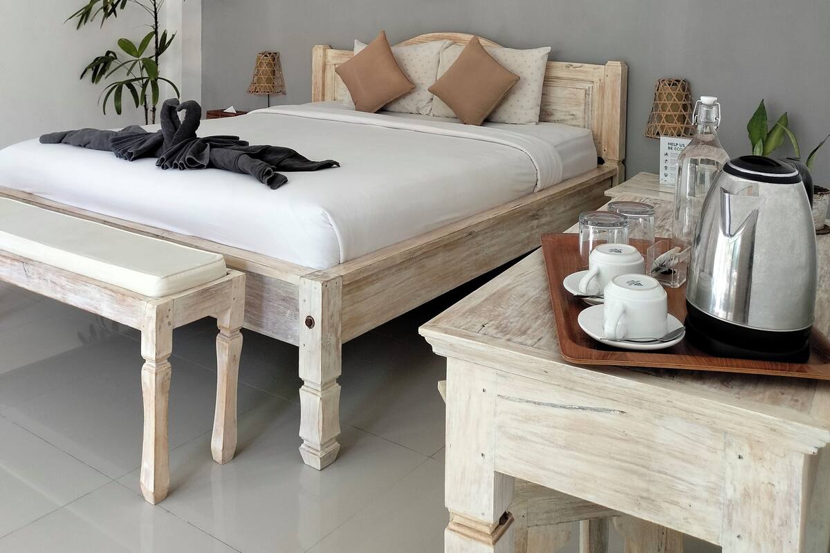 Una cama bien hecha con edredón blanco y dos almohadas marrones, una mesita de noche con una bandeja que contiene un hervidor, tazas y una tetera, y un aire acondicionado de pared.