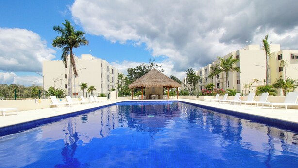 Una piscina con sillas de descanso y una estructura de techo de paja, rodeada de palmeras y edificios.