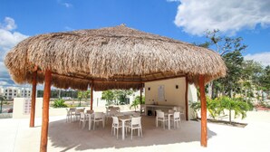 Un área de estar al aire libre con techo de paja, sillas de plástico blanco y una pequeña zona de bar.