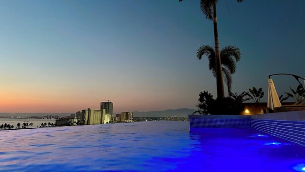 A poolside view with a cityscape in the background, palm trees, and a sunset. - Stunning sunset view from the rooftop infinity pool. 5 min walk to the beach. (Puerto Vallarta)