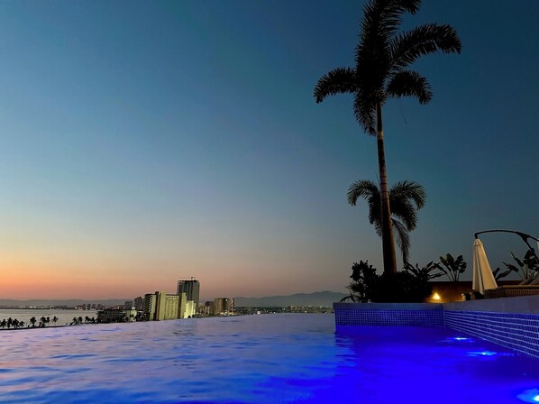 A poolside view with a cityscape in the background, palm trees, and a sunset.