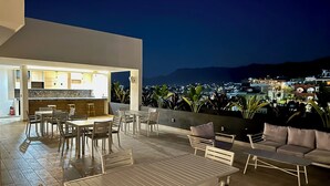 An outdoor patio with a dining area and seating, a kitchen with a bar, and a view of a cityscape at night. - Stunning sunset view from the rooftop infinity pool. 5 min walk to the beach. (Puerto Vallarta)
