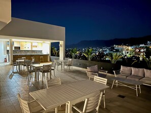 An outdoor patio with a dining area and seating, a kitchen with a bar, and a view of a cityscape at night.