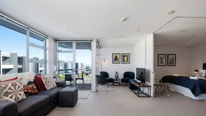 Ein modernes Wohnzimmer mit einer Couch, Sesseln, einem Couchtisch und einem Bett. Große Fenster bieten einen Blick auf die Stadt.