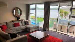 Un salon moderne avec un canapé d’angle gris, des coussins rouges, une table basse en bois et un tapis rouge. De grandes fenêtres offrent une vue sur une étendue d’eau et sur la verdure.