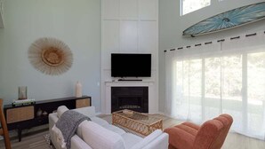 Un salón con un sofá blanco, un sillón marrón, un televisor de pantalla plana sobre una chimenea y un ventanal con cortinas translúcidas.