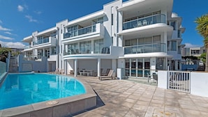 Un edificio moderno de varios pisos con piscina, mobiliario exterior y cielo azul despejado.