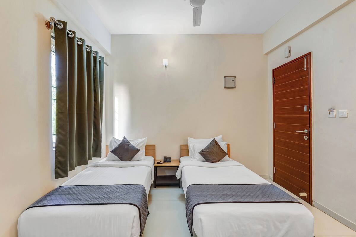 A hotel room with two beds, a wooden door, a nightstand, and a window with curtains.