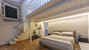 Een slaapkamer met een bed, nachtkastje, bureau, raam en een trap naar een verdieping erboven.